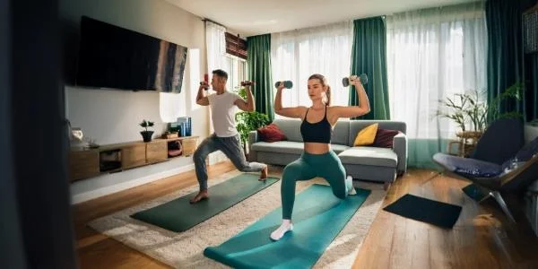 Une femme faisant du yoga et un homme avec des haltères dans un espace workout bien organisé à la maison, avec tapis et accessoires de sport à portée de main.